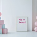 Minimalistic setup with a white wall, a &laquo;&nbsp;Pop to Reveal!&nbsp;&raquo; sign, and pink and blue balloon boxes stacked in a pyramid&nbsp;shape.